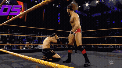 A Wrestler Is Kneeling Down In A Wrestling Ring While Another Wrestler Stands In The Corner GIF