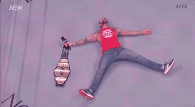 A Wrestler Is Laying On The Floor With A Microphone And A Championship Belt . GIF