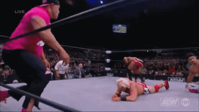A Wrestler Is Laying On The Ground In A Wrestling Ring While A Man In A Pink Shirt Looks On . GIF