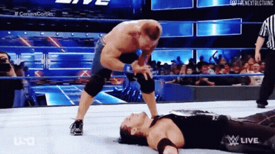 A Wrestler Is Laying On The Ground While Another Wrestler Is Standing On Top Of Him . GIF