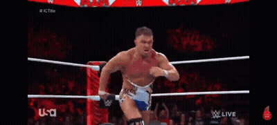A Wrestler Is Standing In The Middle Of A Wrestling Ring While Another Wrestler Is Kneeling Down . GIF