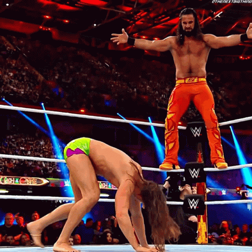A Wrestler Is Standing On A Wrestling Ring Post While Another Wrestler Is On The Ground GIF