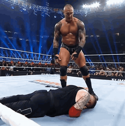 A Wrestler Standing Over Another Wrestler Laying On The Floor GIF