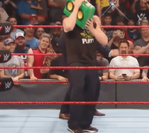 A Wrestler Wearing A Black Shirt That Says Pain Is Holding A Green Box On His Head In A Wrestling Ring GIF