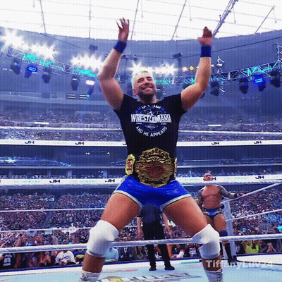 A Wrestler Wearing A Wrestlemania Shirt Stands In A Ring With His Arms In The Air GIF
