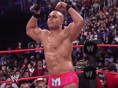 A Wrestler Wearing Pink Shorts With The Letter M On Them Stands In A Wrestling Ring GIF