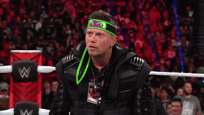 A Wrestler With A Green Headband Is Talking Into A Microphone And The Words What Are On The Screen Behind Him GIF