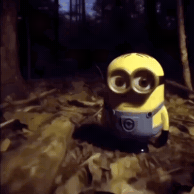 A Yellow Minion With Big Eyes Is Standing In A Dark Forest . GIF