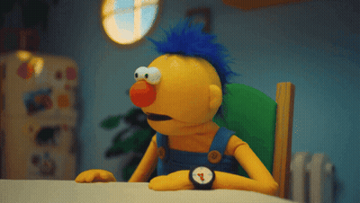 A Yellow Puppet With A Blue Mohawk Sits At A Table GIF