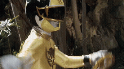 A Yellow Ranger With A Black Helmet And Gloves Is Standing In The Woods . GIF