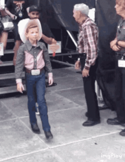 A Young Boy Dressed As A Cowboy Is Walking In Front Of A Group Of People . GIF