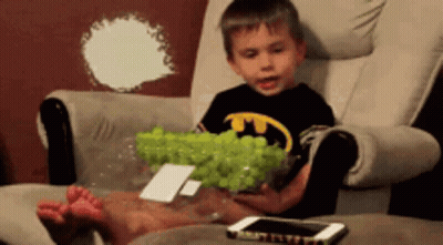 A Young Boy In A Batman Shirt Is Sitting In A Chair Holding A Bowl Of Green Grapes GIF