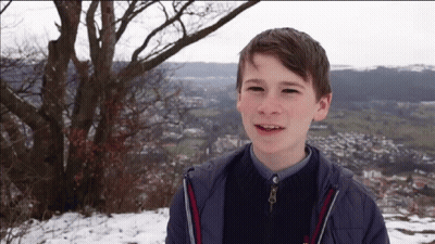 A Young Boy In A Blue Jacket Is Smiling In Front Of A Snowy Landscape GIF