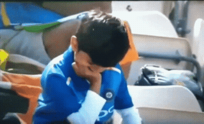 A Young Boy In A Blue Shirt Is Sitting In A Stadium With His Hand On His Face . GIF