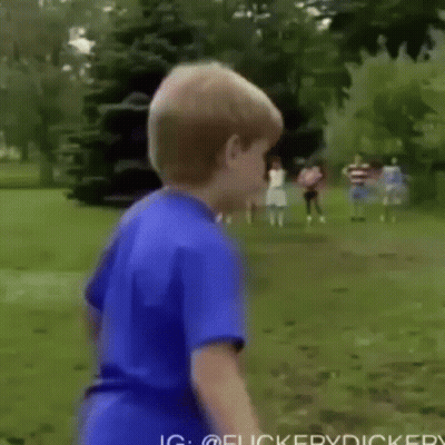 A Young Boy In A Blue Shirt Is Standing In A Field With Trees In The Background . GIF