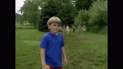A Young Boy In A Blue Shirt Is Standing In A Grassy Field With A Group Of Children In The Background . GIF