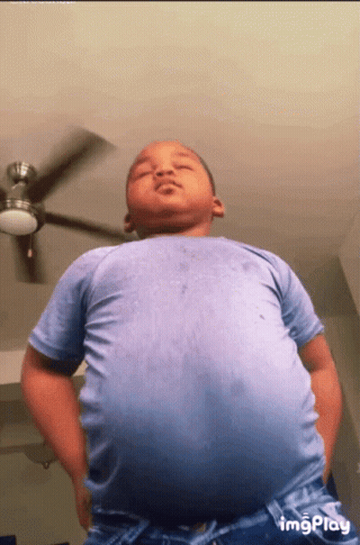 A Young Boy In A Blue Shirt Is Standing Under A Ceiling Fan GIF