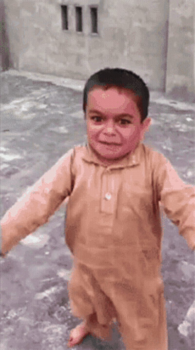 A Young Boy In A Brown Shirt Is Smiling And Walking On A Sidewalk . GIF