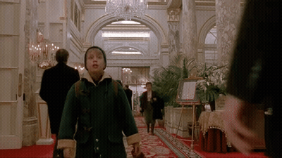 A Young Boy In A Green Coat Is Walking Through A Hallway GIF