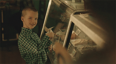 A Young Boy In A Green Plaid Shirt Is Smiling In Front Of A Display Case Of Food GIF