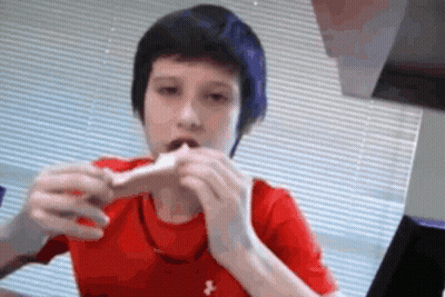 A Young Boy In A Red Under Armour Shirt Is Eating A Sandwich GIF