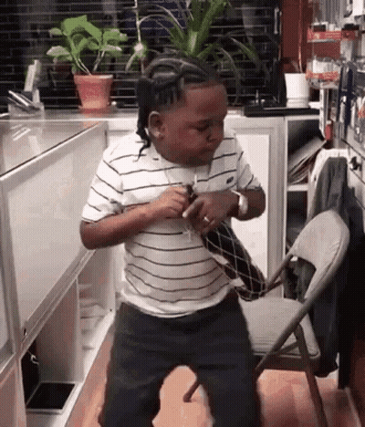 A Young Boy In A Striped Shirt Is Dancing In A Kitchen Next To A Chair . GIF