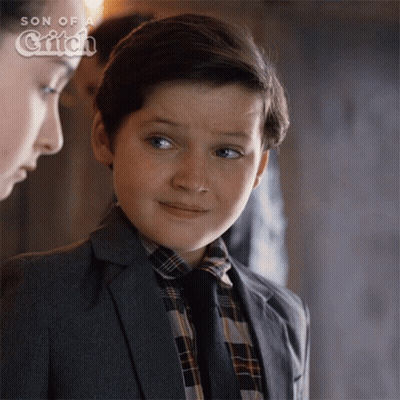 A Young Boy In A Suit And Tie Stands Next To A Woman In A Son Of A Witch Ad GIF