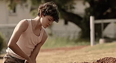 A Young Boy In A Tank Top Is Digging In The Dirt In A Field . GIF
