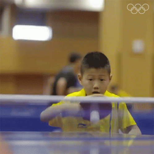 A Young Boy In A Yellow Shirt Is Playing Ping Pong With The Olympic Rings In The Background GIF