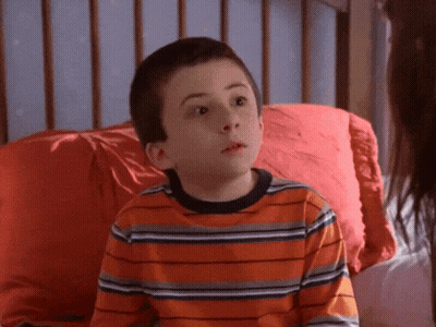 A Young Boy In An Orange Striped Shirt Sits On A Bed With A Red Pillow GIF