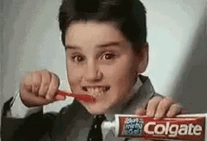 A Young Boy Is Brushing His Teeth While Holding A Tube Of Colgate . GIF