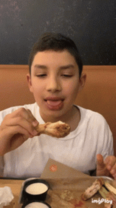 A Young Boy Is Eating A Chicken Wing With His Tongue Hanging Out GIF