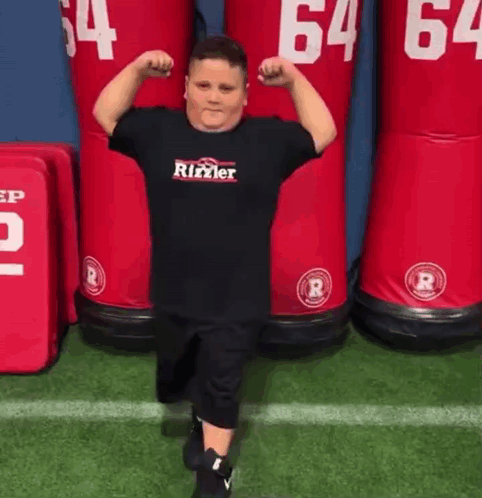 A Young Boy Is Flexing His Muscles On A Football Field In Front Of A Row Of Red Dummies . GIF