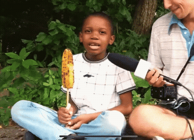 A Young Boy Is Holding A Corn On The Cob And Talking Into A Microphone GIF