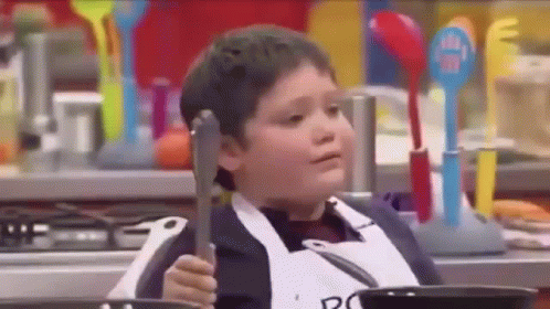 A Young Boy Is Holding A Pair Of Tongs While Sitting In Front Of A Pan . GIF