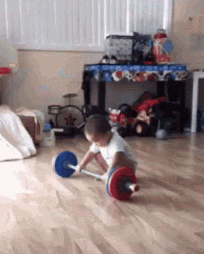 A Young Boy Is Playing With A Barbell On The Floor GIF