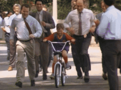 A Young Boy Is Riding A Bike With A Group Of Men Following Behind Him GIF