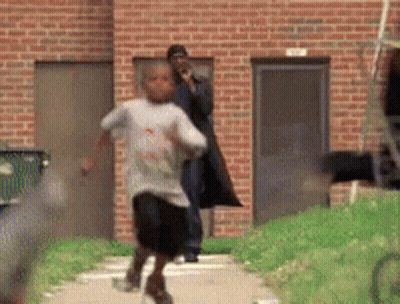a young boy is running down a sidewalk in front of a brick building while a man watches .