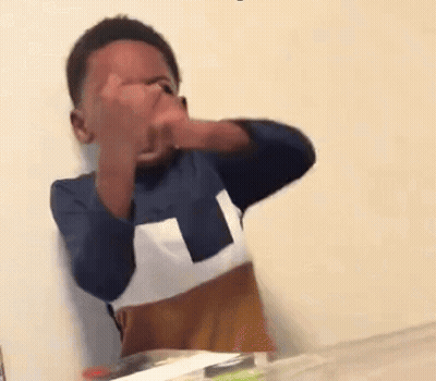 A Young Boy Is Sitting At A Table Covering His Face With His Hands . GIF