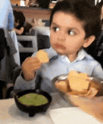 A Young Boy Is Sitting At A Table Eating Chips And Guacamole . GIF