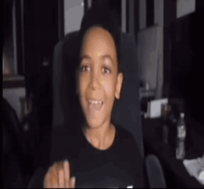 A Young Boy Is Sitting In A Chair With His Mouth Open And Pointing . GIF