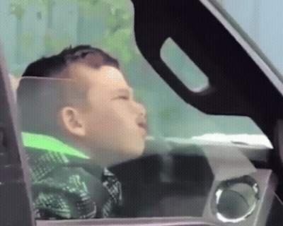 A Young Boy Is Sitting In The Driver 'S Seat Of A Car Looking Out The Window GIF
