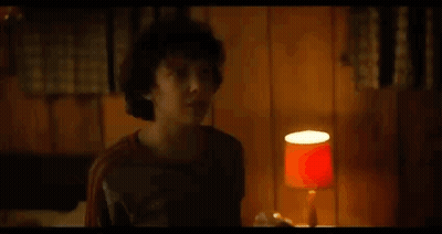 A Young Boy Is Standing In A Room With A Lamp And A Wooden Wall . GIF