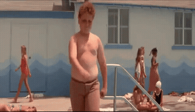 A Young Boy Is Standing In Front Of A Swimming Pool While A Group Of Children Are Playing In The Water . GIF