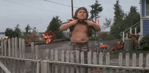 A Young Boy Is Standing In Front Of A Wooden Fence And Looking Over It . GIF