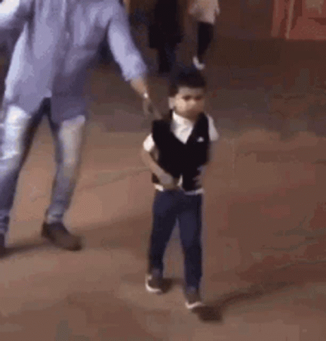 A Young Boy Is Standing On A Tiled Floor With His Hands In His Pockets And A Man Walking Behind Him . GIF