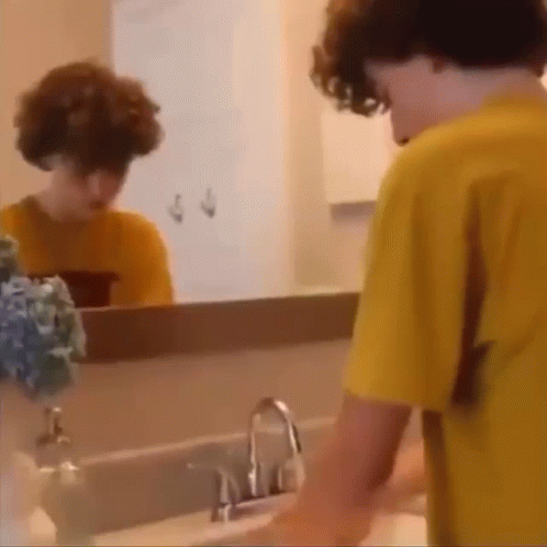 A Young Boy Is Washing His Hands In A Bathroom Sink While Looking At Himself In The Mirror . GIF