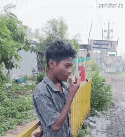 A Young Boy Smoking A Cigarette In Front Of A Yellow Fence With The Website Bavishni.in Written On The Bottom GIF
