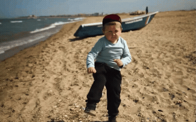 A Young Boy Stands On A Beach With A Boat In The Background GIF
