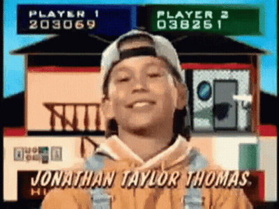 A Young Boy Wearing A Baseball Cap And Overalls Is Smiling In Front Of A House . GIF